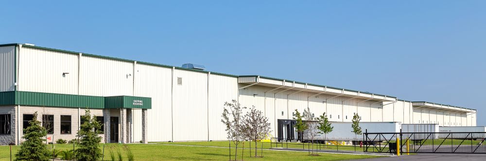 Light-colored commercial warehouse building with green accent panels, office entrance, loading docks, and landscaped lawn under a clear blue sky.